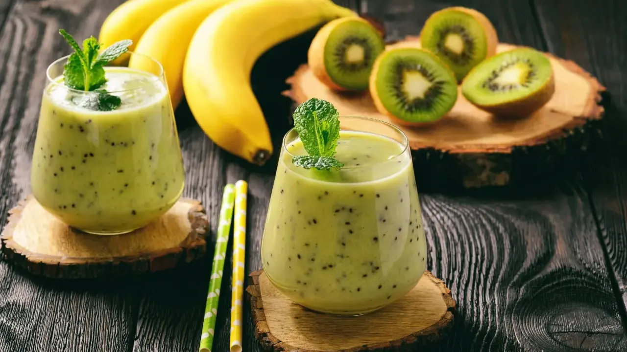 Deux verres de smoothie banane kiwi, décorés de menthe, avec des bananes et des kiwis coupés en arrière-plan.