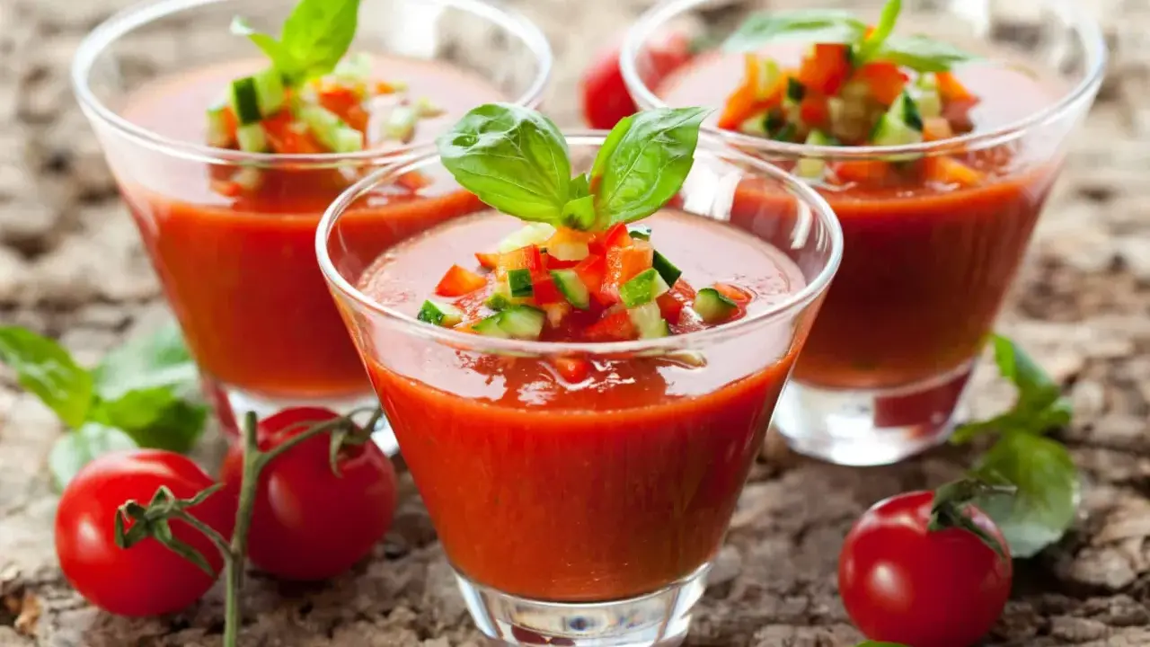 Trois verres de gaspacho tomate, garni de dés de légumes et de basilic, sur fond de bois.