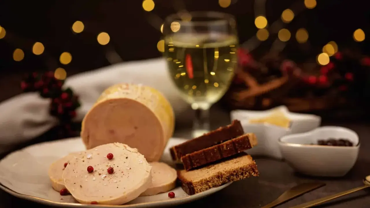 Tranche de foie gras sur une assiette, accompagnée de pain d'épices et d'un verre de vin blanc. Ambiance festive avec des lumières scintillantes.