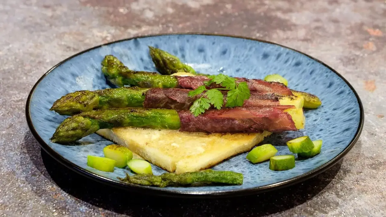 Asperge à la poêle enroulée de viande séchée, servie sur du pain grillé, garnie de persil frais et de morceaux d'asperges.