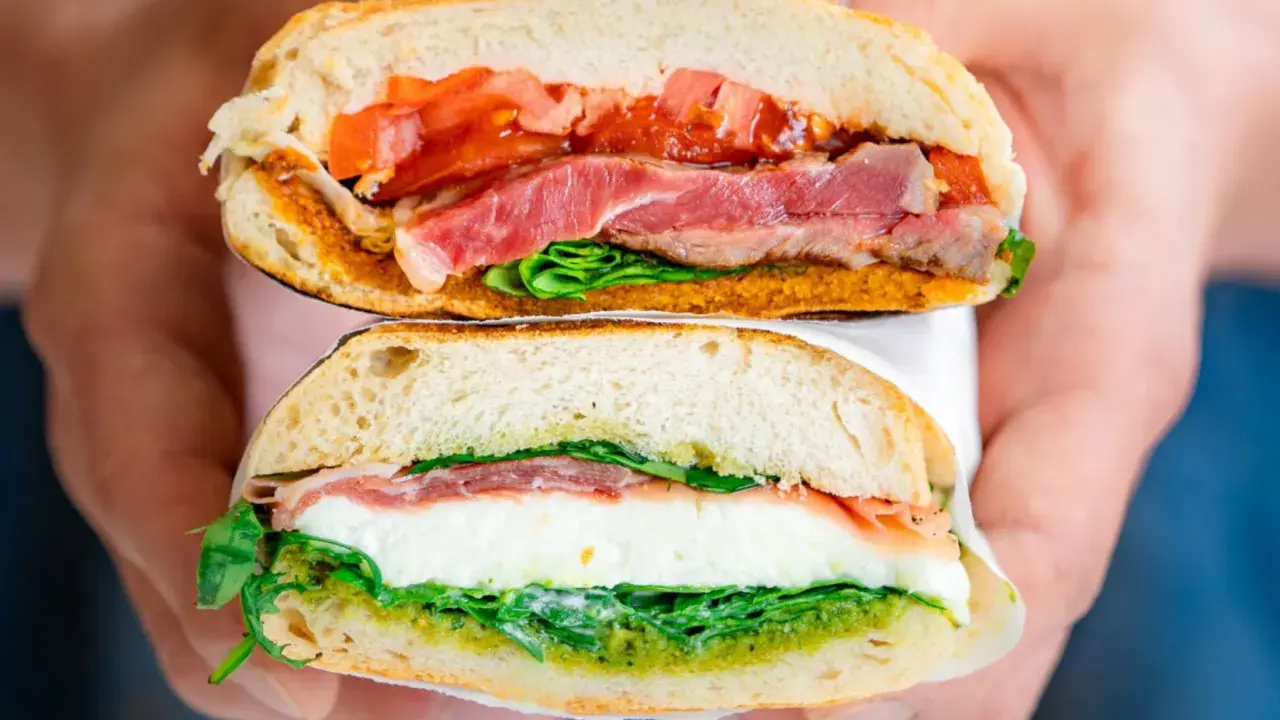 Deux moitiés d'un délicieux sandwich italien, garni de jambon, de tomates, de mozzarella et de pesto, sont tenues par des mains.