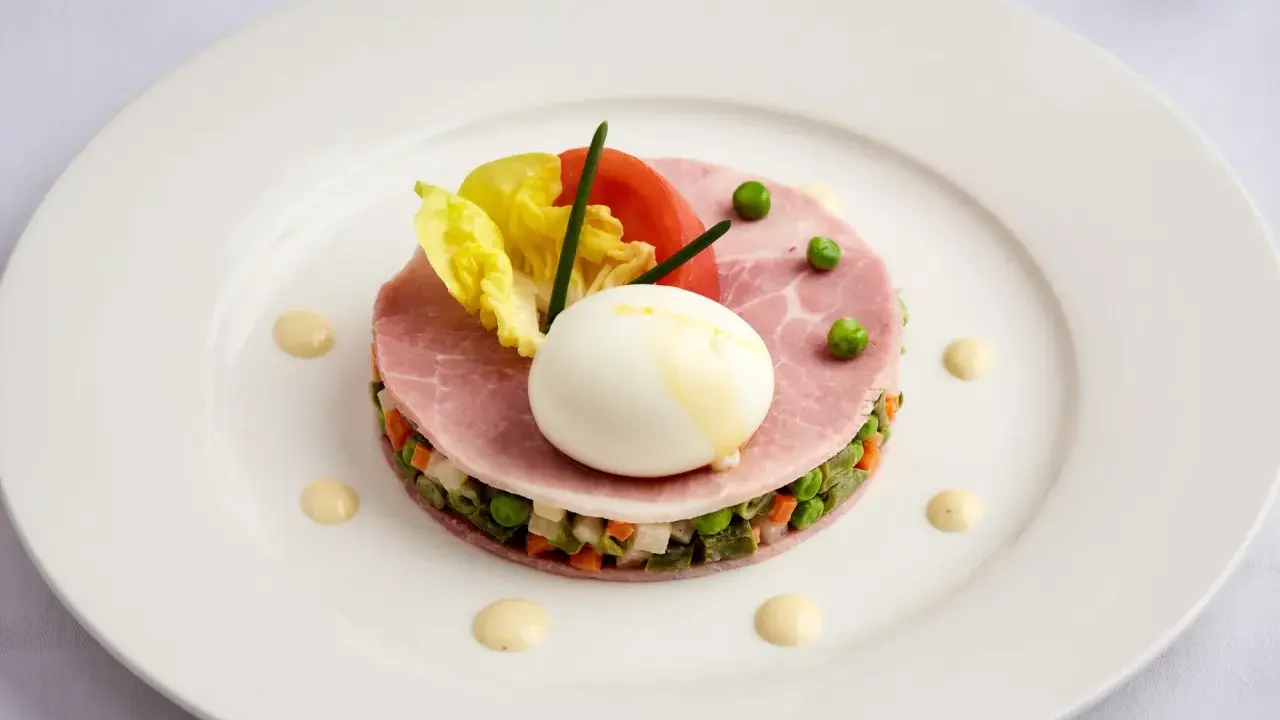 Une assiette blanche présente une délicieuse macédoine de légumes, garnie de jambon, d'un œuf mollet, de tomates et de laitue.