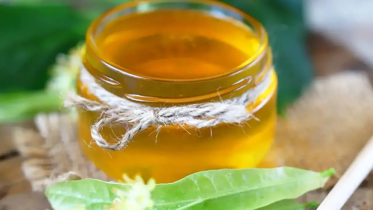 Un pot de sirop de tilleul doré, noué de cordelette, entouré de feuilles vertes et de fleurs de tilleul.