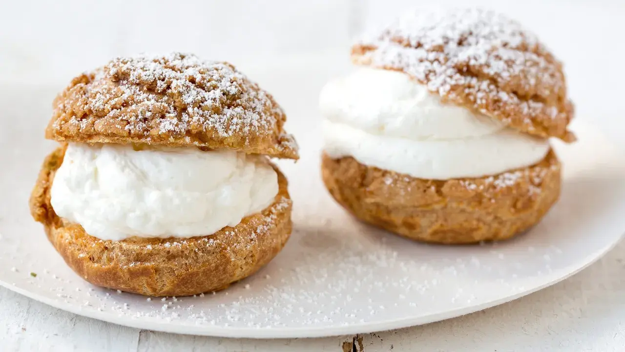 Deux choux à la crème généreusement garnis de crème fouettée, saupoudrés de sucre glace sur une assiette blanche.