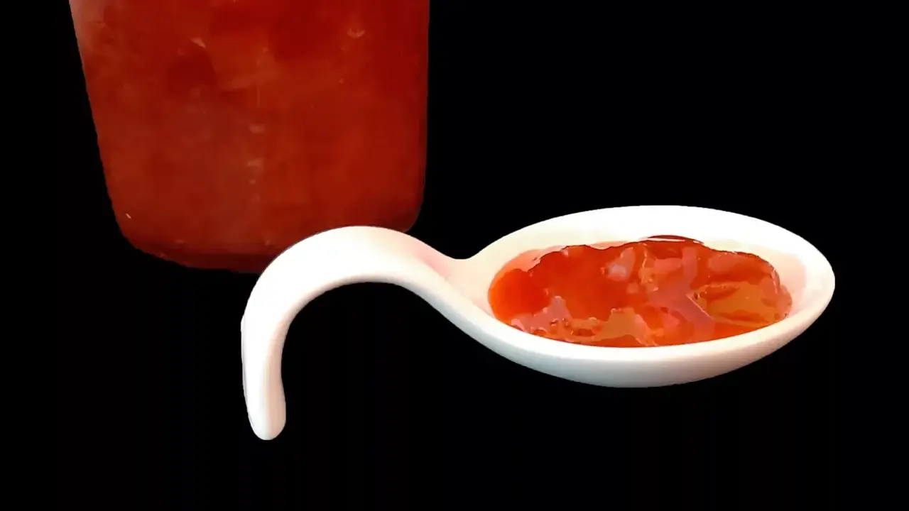 Une cuillère blanche présente une délicieuse confiture fraise rhubarbe, avec un pot en arrière-plan.