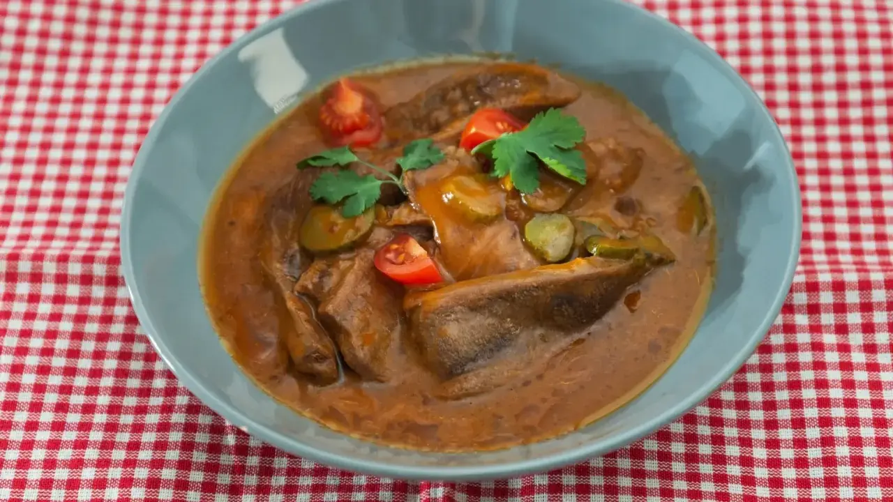 Langue de bœuf en sauce piquante, garnie de cornichons, tomates cerises et persil, servie dans un bol bleu sur une nappe à carreaux rouges et blancs.