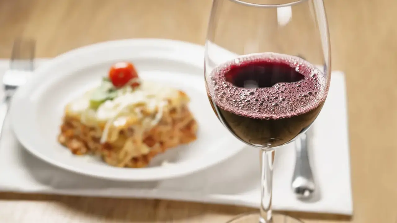 Un verre de vin rouge pétillant à côté d'une portion de lasagnes appétissante. La question se pose : quel vin avec des lasagnes pour un accord parfait ?