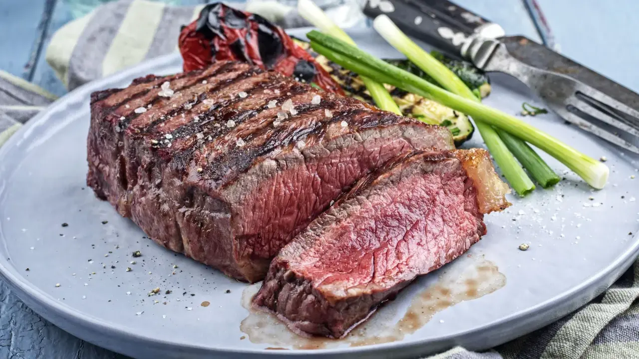 Un savoureux rumsteck de boeuf grillé, coupé en tranches, servi avec des légumes rôtis et des oignons verts sur une assiette grise.