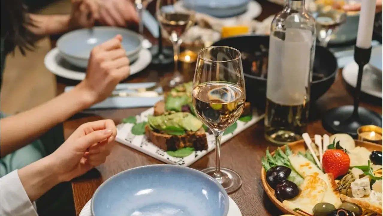 Un verre de vin blanc beurré, des toasts à l'avocat et un plateau de fromages et fruits sur une table dressée pour un dîner convivial.