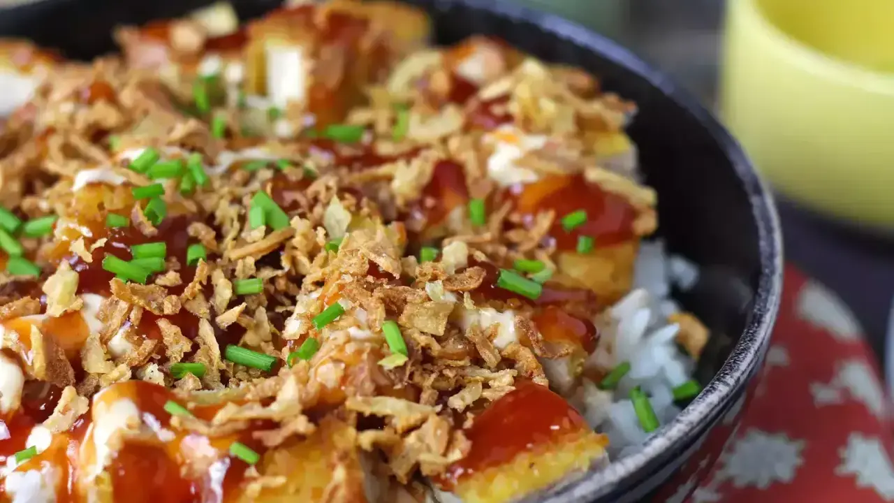 Un bol de riz garni de morceaux de crispy de poulet, nappé de sauce et parsemé d'oignons frits et de ciboule.