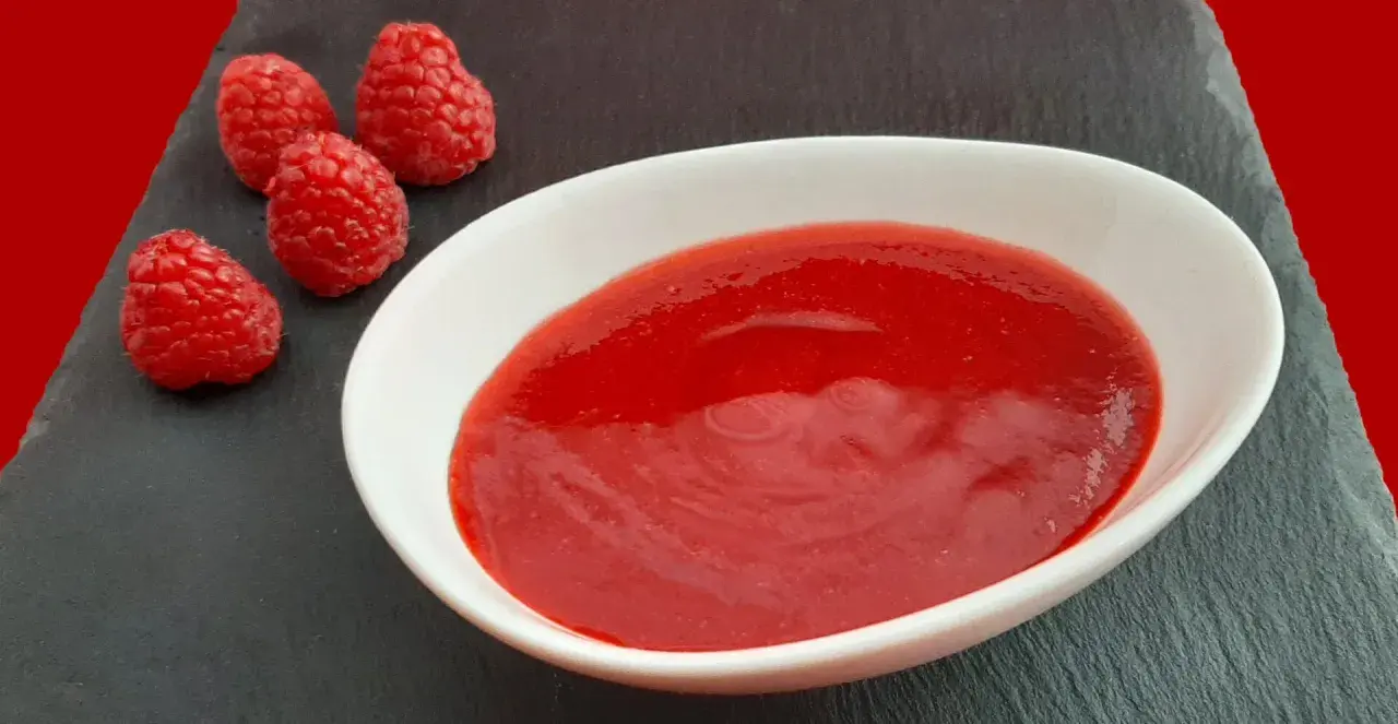 Un bol blanc contient une **purée de framboise** onctueuse, accompagnée de quelques framboises fraîches sur une ardoise.