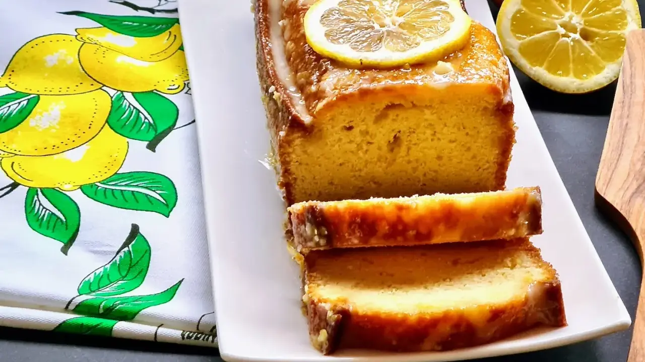 Gâteau moelleux au citron, glacé et garni de rondelles de citron. Ce délice, un gâteau huile d'olive, est servi sur une assiette blanche avec un torchon orné de citrons.