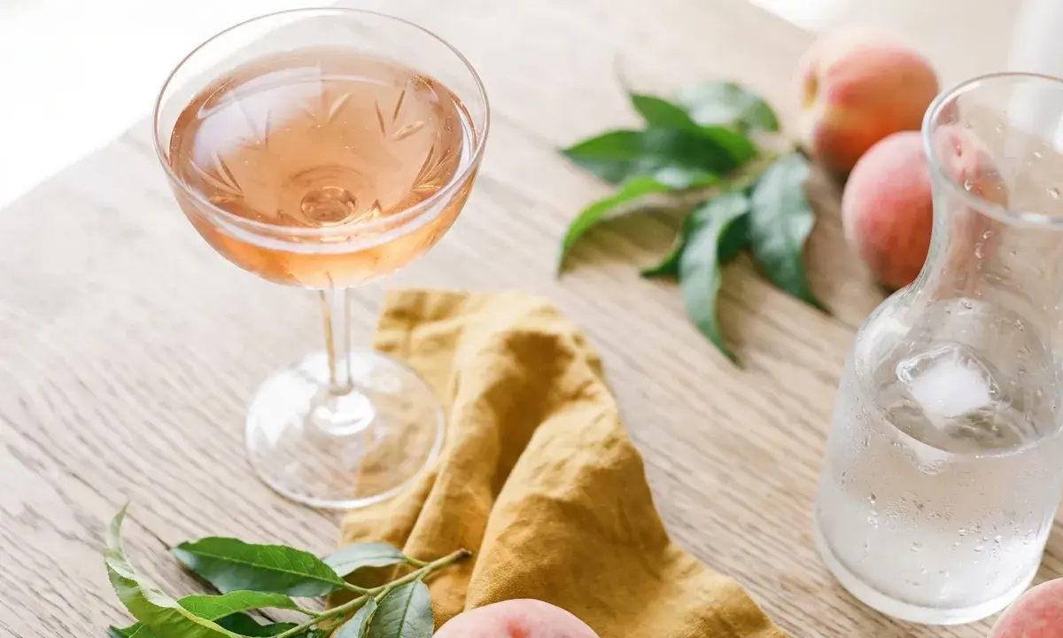 Un verre de vin de pêche rosé, des pêches fraîches et une carafe d'eau glacée sur une table en bois.