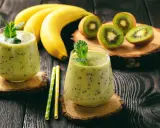 Deux verres de smoothie banane kiwi, décorés de menthe, avec des bananes et des kiwis coupés en arrière-plan.