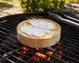 Un camembert au barbecue fond sur les braises. Des pinces à barbecue sont posées à côté, prêtes à servir ce délice crémeux.