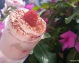 Un tiramisu framboise rapide et facile, garni d'une framboise fraîche et de cacao, avec des fleurs roses en arrière-plan.
