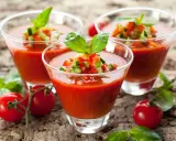 Trois verres de gaspacho tomate, garni de dés de légumes et de basilic, sur fond de bois.
