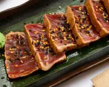 Tranches de tataki de boeuf, poêlé et saupoudré de graines de sésame noires et dorées, servi avec une touche de wasabi sur une feuille verte.