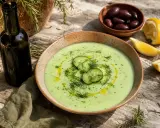 Un bol de soupe de concombre froide, garni de rondelles de concombre et d'aneth, avec des olives, du citron et une bouteille d'huile d'olive.