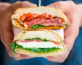 Deux moitiés d'un délicieux sandwich italien, garni de jambon, de tomates, de mozzarella et de pesto, sont tenues par des mains.