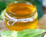 Un pot de sirop de tilleul doré, noué de cordelette, entouré de feuilles vertes et de fleurs de tilleul.
