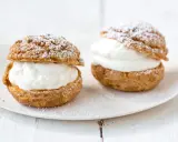 Deux choux à la crème généreusement garnis de crème fouettée, saupoudrés de sucre glace sur une assiette blanche.