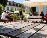Verre de vin blanc sec ou moelleux sur une table en bois, avec des gens attablés en arrière-plan dans un jardin ensoleillé.