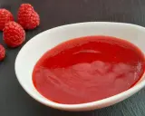 Un bol blanc contient une **purée de framboise** onctueuse, accompagnée de quelques framboises fraîches sur une ardoise.