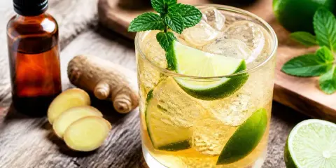 Un verre de mocktail ginger beer rafraîchissant, garni de menthe et de quartiers de citron vert, avec du gingembre frais et une fiole d'extrait à proximité.