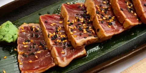 Tranches de tataki de boeuf, poêlé et saupoudré de graines de sésame noires et dorées, servi avec une touche de wasabi sur une feuille verte.