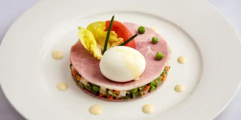 Une assiette blanche présente une délicieuse macédoine de légumes, garnie de jambon, d'un œuf mollet, de tomates et de laitue.