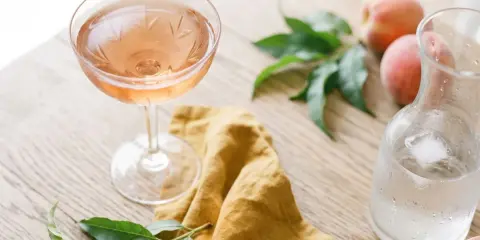 Un verre de vin de pêche rosé, des pêches fraîches et une carafe d'eau glacée sur une table en bois.