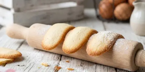 Des biscuits langue de chat dorés, saupoudrés de sucre glace, reposent sur un rouleau à pâtisserie en bois.