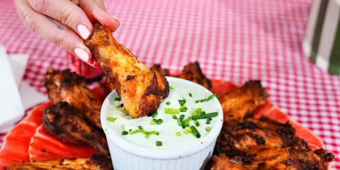 Une main trempe une aile de poulet buffalo dans une sauce crémeuse. Les ailes sont disposées sur une assiette rouge, sur une nappe à carreaux.