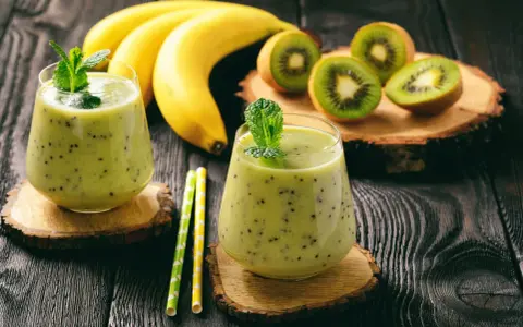 Deux verres de smoothie banane kiwi, décorés de menthe, avec des bananes et des kiwis coupés en arrière-plan.
