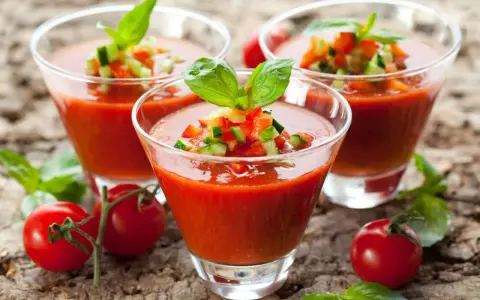 Trois verres de gaspacho tomate, garni de dés de légumes et de basilic, sur fond de bois.