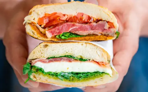 Deux moitiés d'un délicieux sandwich italien, garni de jambon, de tomates, de mozzarella et de pesto, sont tenues par des mains.