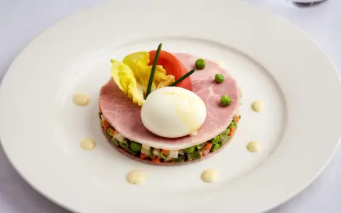 Une assiette blanche présente une délicieuse macédoine de légumes, garnie de jambon, d'un œuf mollet, de tomates et de laitue.