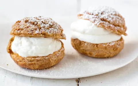 Deux choux à la crème généreusement garnis de crème fouettée, saupoudrés de sucre glace sur une assiette blanche.