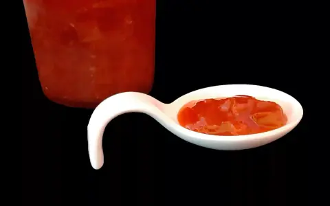Une cuillère blanche présente une délicieuse confiture fraise rhubarbe, avec un pot en arrière-plan.