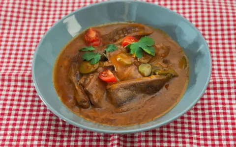 Langue de bœuf en sauce piquante, garnie de cornichons, tomates cerises et persil, servie dans un bol bleu sur une nappe à carreaux rouges et blancs.