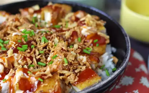 Un bol de riz garni de morceaux de crispy de poulet, nappé de sauce et parsemé d'oignons frits et de ciboule.