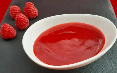 Un bol blanc contient une **purée de framboise** onctueuse, accompagnée de quelques framboises fraîches sur une ardoise.