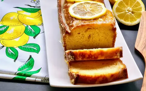 Gâteau moelleux au citron, glacé et garni de rondelles de citron. Ce délice, un gâteau huile d'olive, est servi sur une assiette blanche avec un torchon orné de citrons.