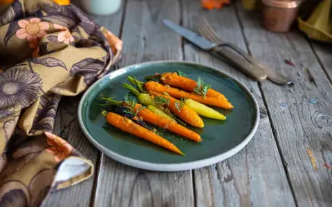Carottes glacées au miel, colorées et appétissantes, garnissent une assiette sur une table en bois rustique. Une serviette à motifs floraux complète la scène.