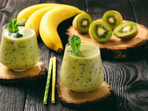 Deux verres de smoothie banane kiwi, décorés de menthe, avec des bananes et des kiwis coupés en arrière-plan.