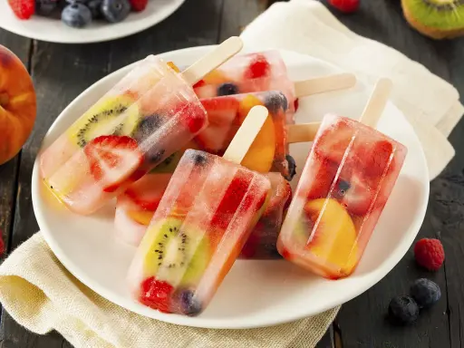 Plusieurs bâtonnets de glace à l'eau fruitée, remplis de fraises, kiwis, pêches et myrtilles, sur une assiette blanche.