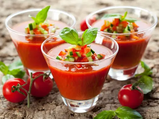 Trois verres de gaspacho tomate, garni de dés de légumes et de basilic, sur fond de bois.