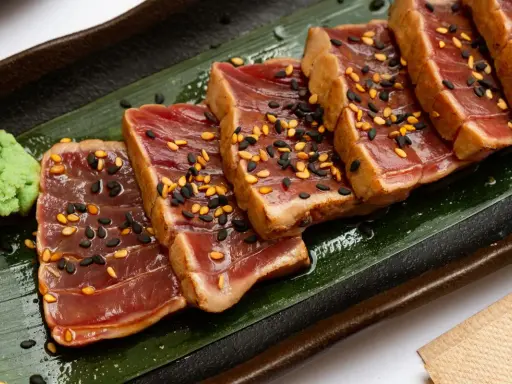 Tranches de tataki de boeuf, poêlé et saupoudré de graines de sésame noires et dorées, servi avec une touche de wasabi sur une feuille verte.
