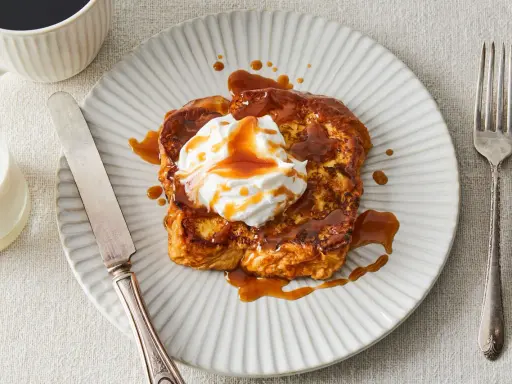 Pain perdu gourmand nappé d'une sauce caramel au miso, garni de crème fouettée. Servi avec une tasse de café et un verre de lait.